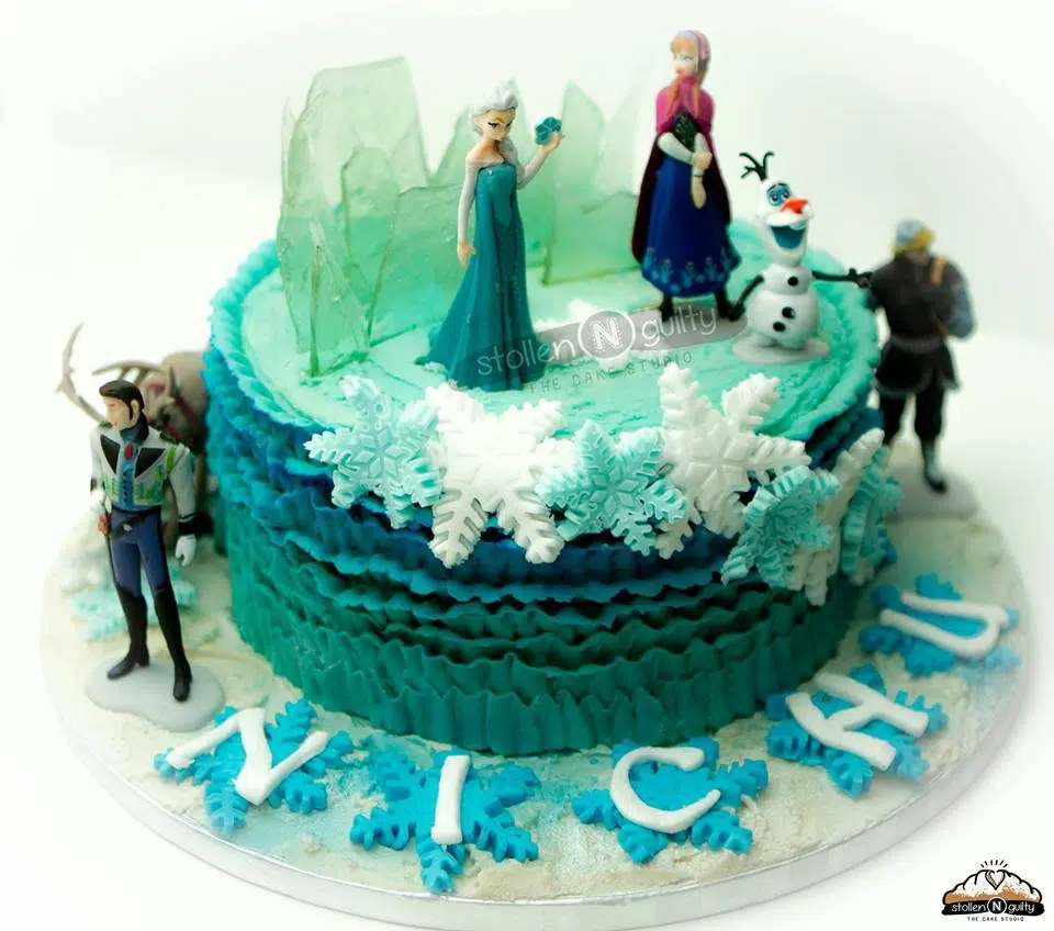 A round cake decorated with buttercream frosting, edible sugar glass and snowflakes, and figurines of all the main characters from Frozen. Stollen N Guilty. Source