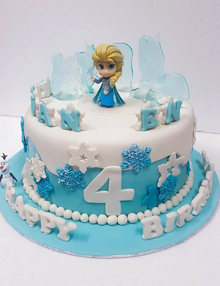 A tall round cake decorated with white and blue fondant cutouts, sugar glass and figurines of Frozen characters. Little Sprinkles. Source