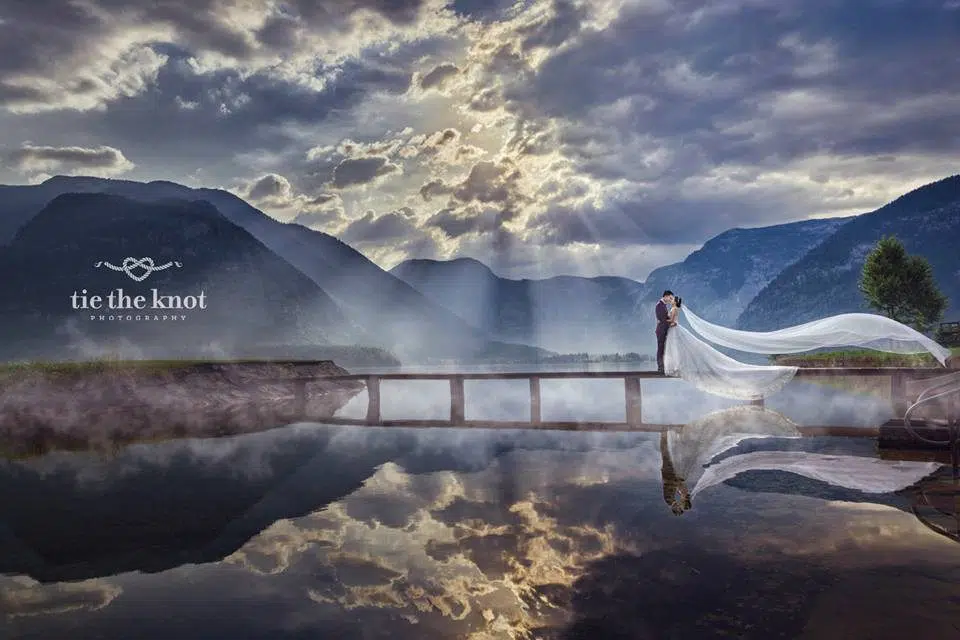 Pre-wedding photograph of couple on a footbridge across a lake in Halstatt Austria. By Tie The Knot Photography. Get quotes for your destination wedding at Recommend.my